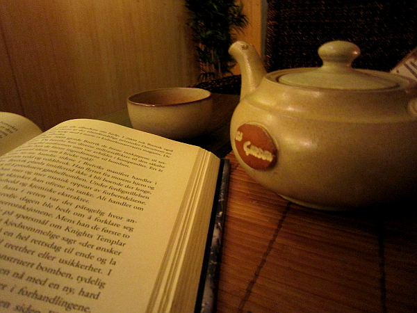 A book is open on a table, there's a tea cup in the back, and a teapot to the right.