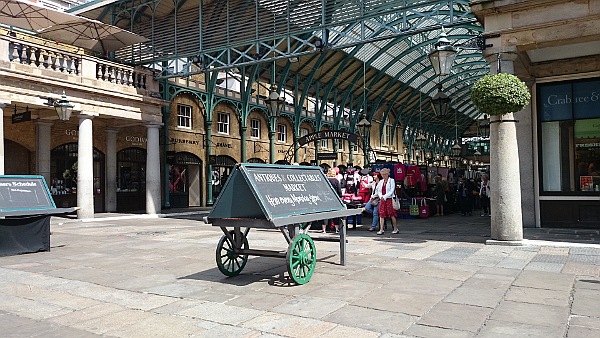 Inside a market hall. A sign with wheels. You can see there's white writing on blackboard on the sign, but it's not readable.