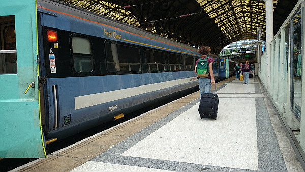 A blue train by the platform, one door is open. On the carriage "First class" is writte with orange letters. On the platform you can see the back of a person walking down, dragging a small suitcase with wheels behind them. They also have a gree rucksack on their back.