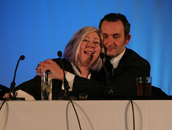 Lady Elsie being hugged by Herr Döktor, both sitting behind a table with a white cloth and microphones. 