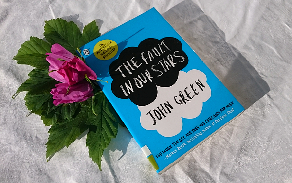 The book "The fault of our stars" on a white table cloth, pink roses with green leaves are tucked in under the top left corner.