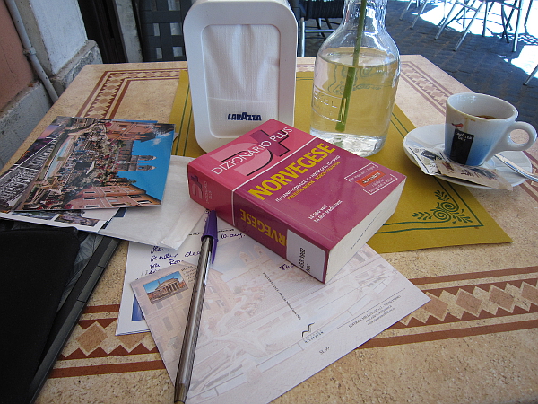 A table with postcards, a cup of coffee, and on top of the postcards there's a Norwegian - Italian, Italian - Norwegian dictionary.