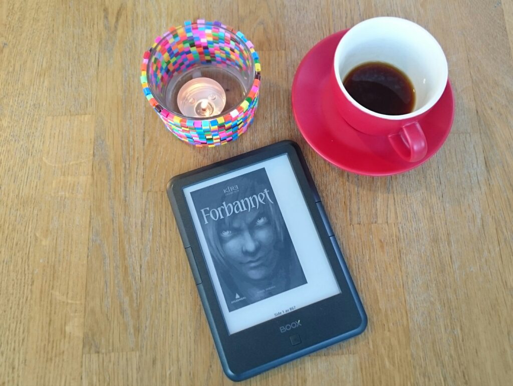 An eReader is laying on a table. Behind it is a lit candle, and a cup of coffee.