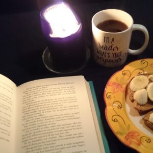 An open book to the left, a plate with breadwith spred can partly be seen on the right. Behind them there's a lit black candle, and a cup of coffee. 