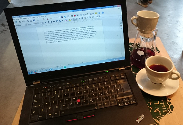 An open laptop computer, showing a text document. To the right there's a carafle of coffee, as well as a cup of coffee.