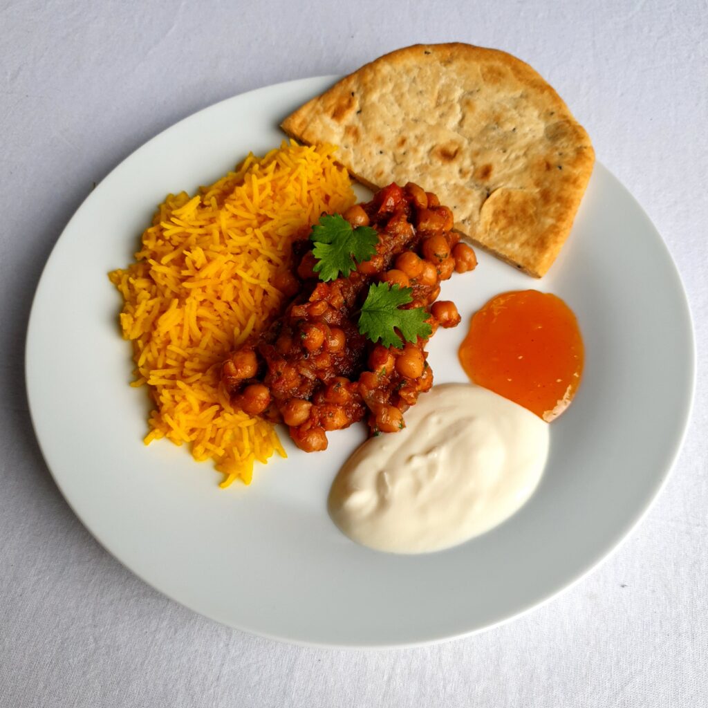 A plate with chana masala, yellow rice, soy yoghurt, mango chutney, and naan.