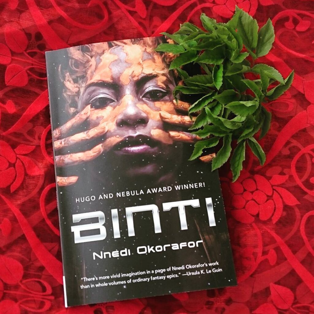 The book "Binti" laying on a red sheer fabric with swirl pattern, there are some green leaves tucked in under it to the top right.