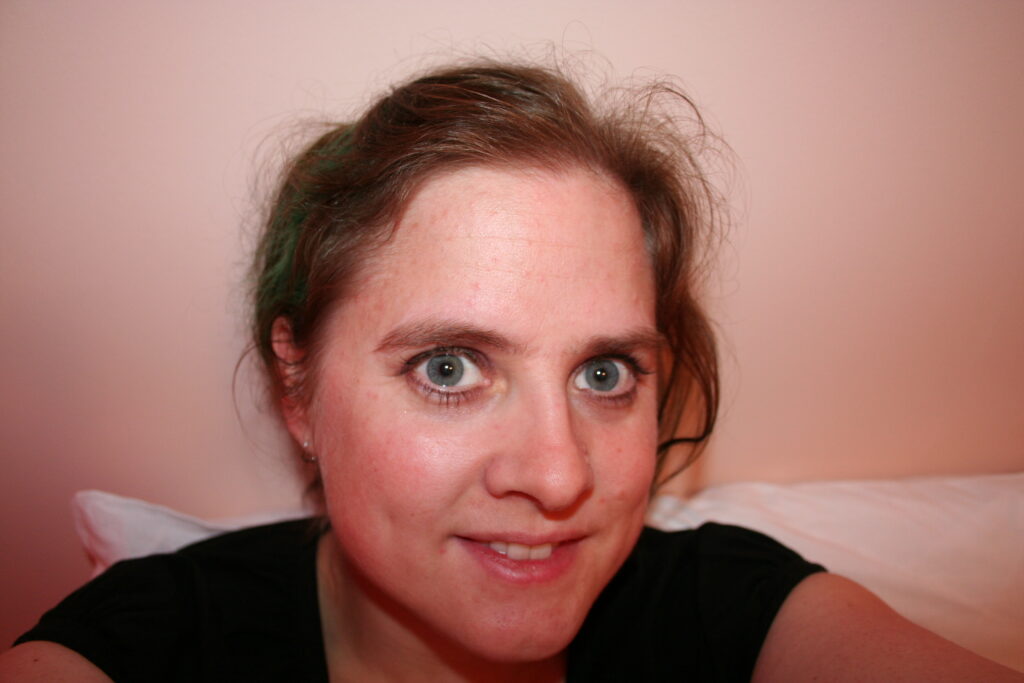 A woman in a hotel room with a black t-shirt. Her hair is blonde with green parts.