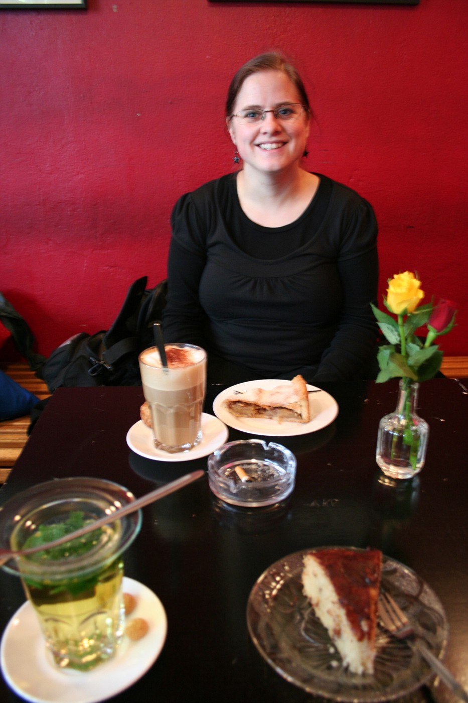 A woman siting behind a table. The wall behind her is red. On the table in front of her there are coffee and cakes.