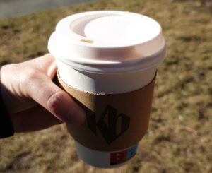 A hand holding a papercup of takeaway coffee outside. Brown grass in the background, after the snow has melted.