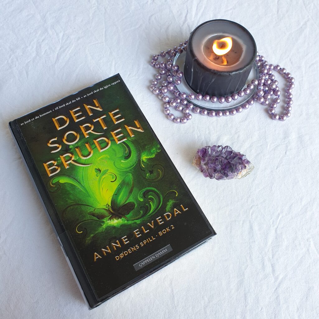 The book «Den sorte bruden» on a table with a white tablecloth, to the right there's and amethyst, as well as a lit. black candle with a purple pearl necklace around it.