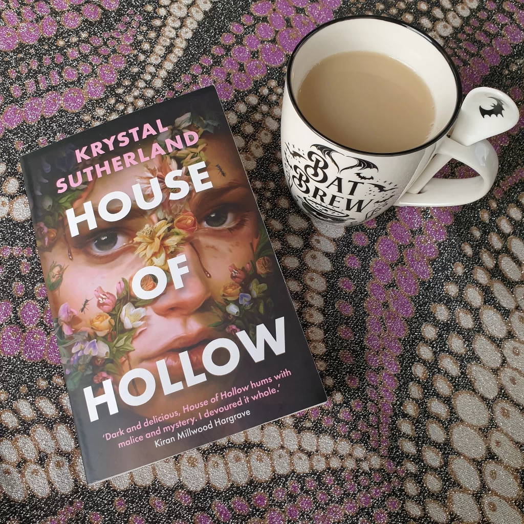 The book «House of Hollow» on a table covered in a fabric with sparkly purples, to the right there's a mug of tea, with the text "Bat brew" on it.