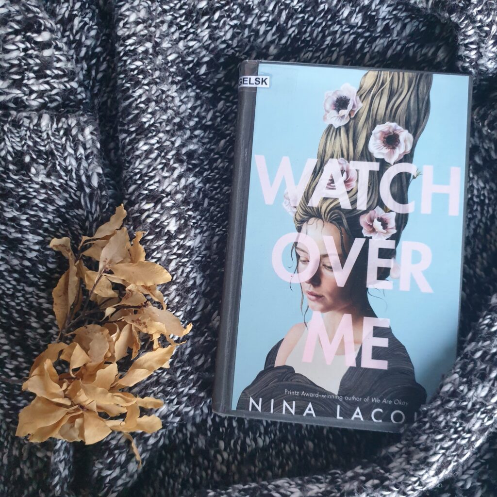 The book «Watch Over Me» in tucked into a grey cardigan. To the left there are some beige, dried leaves.