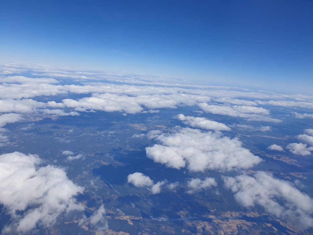 View from an airplane. You can see fields and woods unddr a scattered layer of clouds.