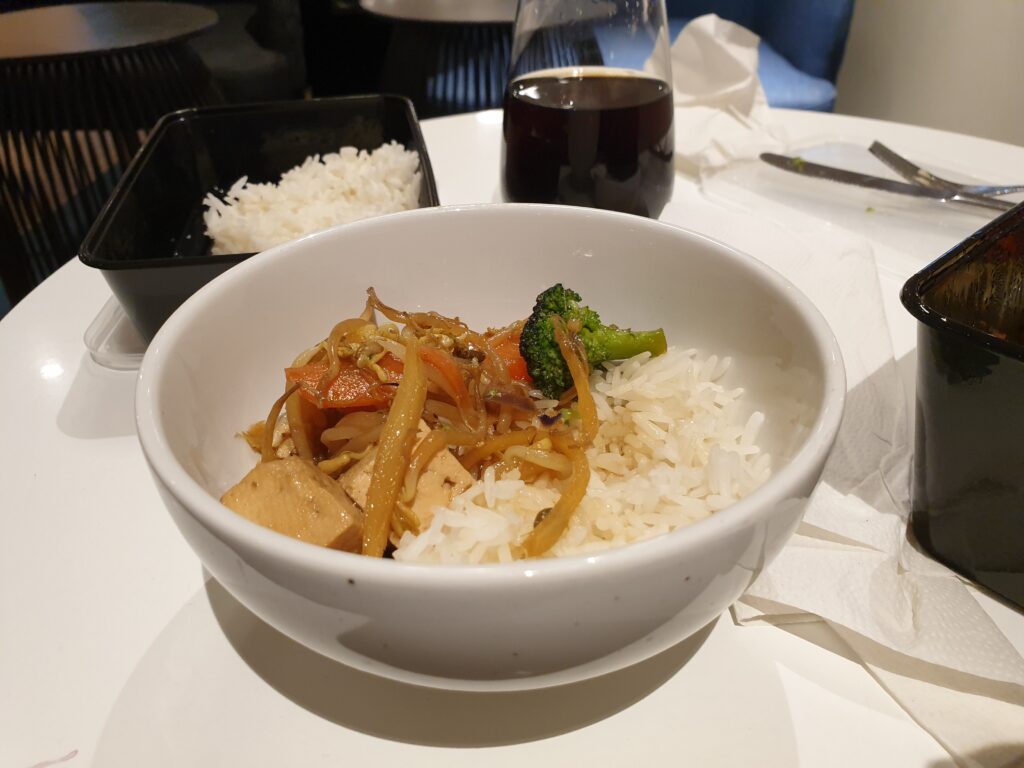 A white bowl on a table. In the bowl there are rice, and wokked vegetables and tofu.
