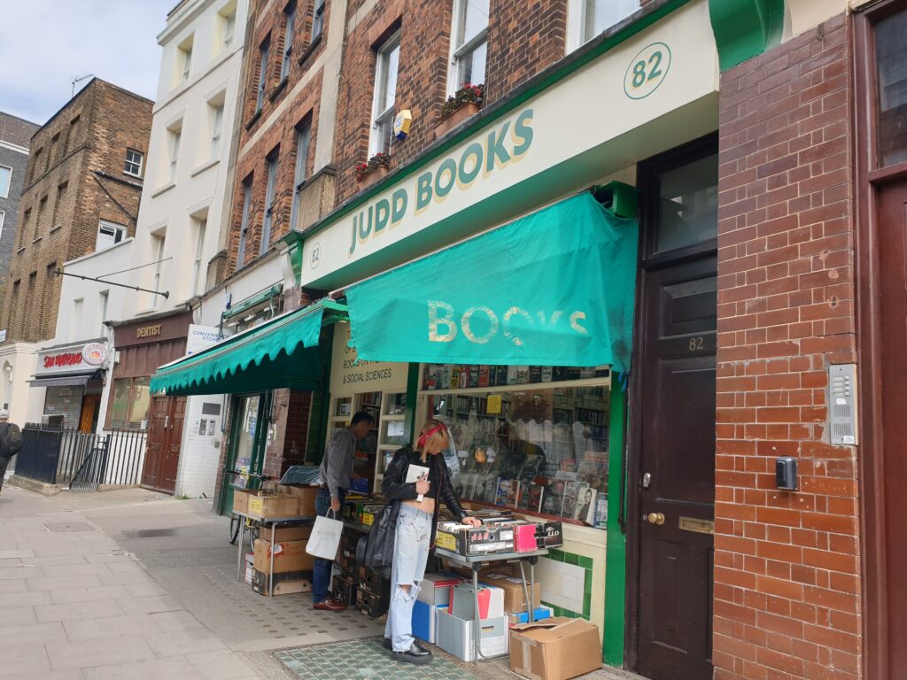 A book shop front, with boxes of books outside. A couple of people can be seen browsing the boxes.