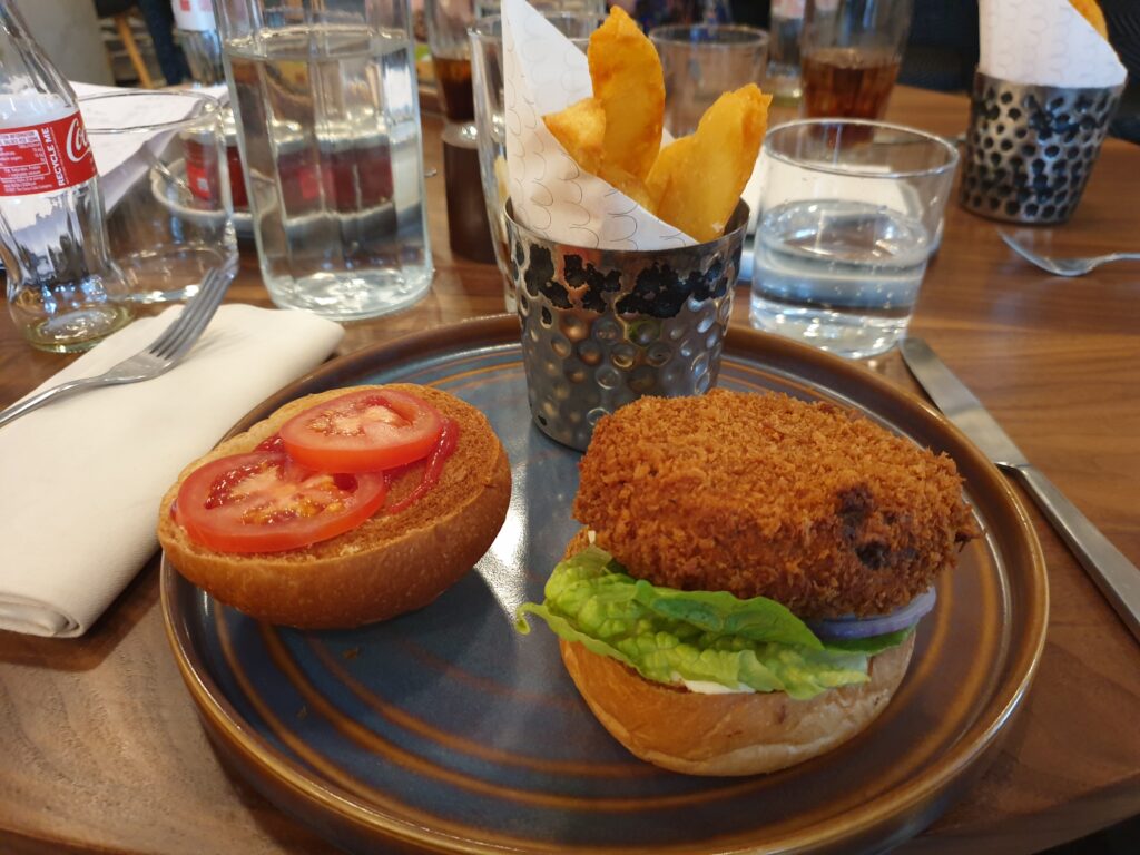 A plate with a burger on a bun half, the other bun half beside it. Also a bowl of chips on it. Behind it are glasses of water.