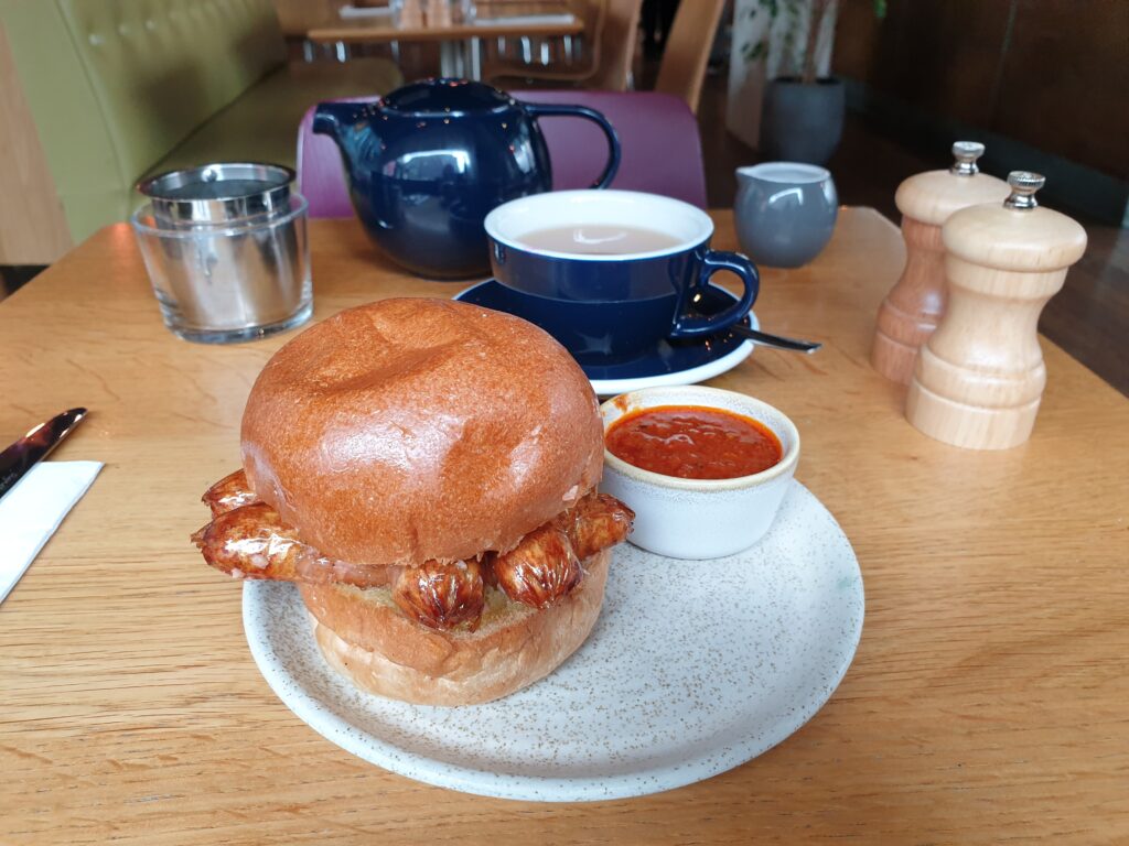 In the front a plate with a bap with sausages and a pot of red chili chutney. Behind it is a mug og tea, as well as a pot of tea.