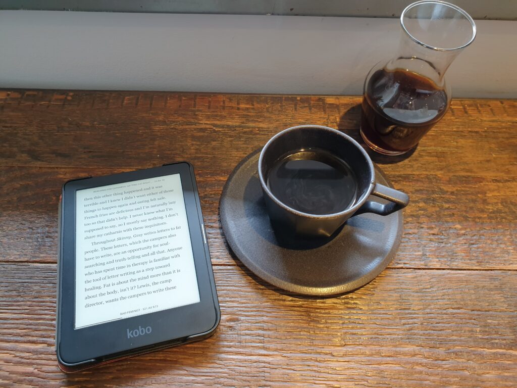 An ereader with a page open to the left, to the right a cup of coffee. A carafe of coffee behind the mug.