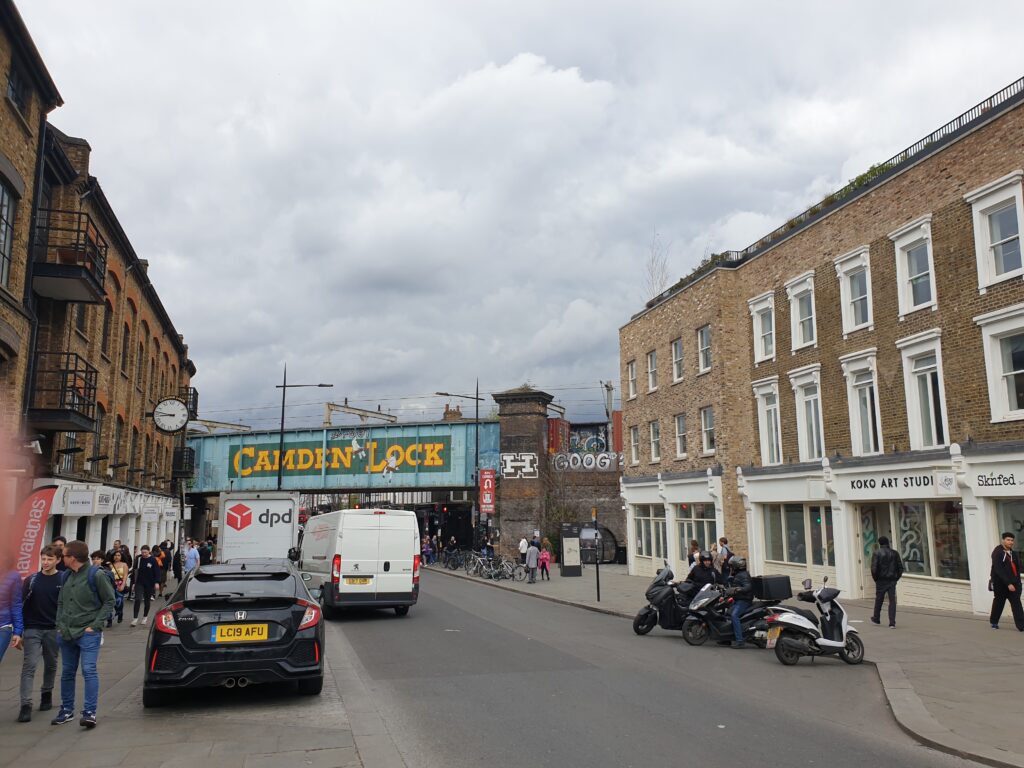 A street eith an enclosed bridge over it- The bridge says "Camden Lock". On each side of the street you can see brick buildings.