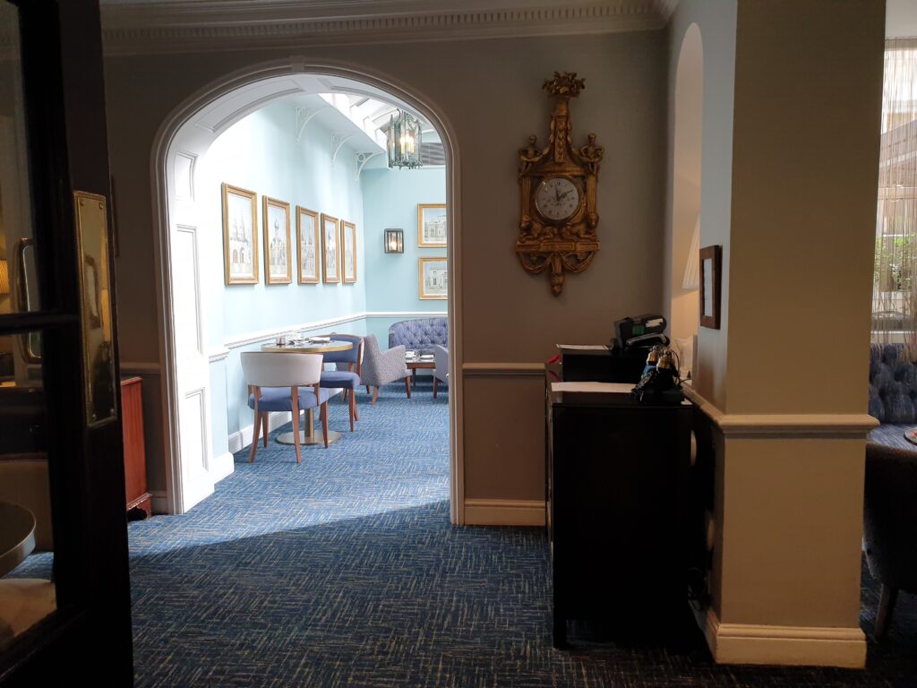 You can see into a sunlit room, with tables and comfortable chairs. The room is blue. On the wall inside the room you can see drawings. To the right of the arch leading into the room, a gold clock is hanging on the wall.