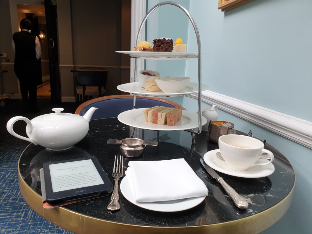 A table with a pot of tea to the left. In the middle is a stand with sandiches, scones, and cakes. To the right there's a cup of tea. In fron there is a plate with a white napkin, a knife and fork at each side. To the left of the plate there's an e-reader, with a page open.