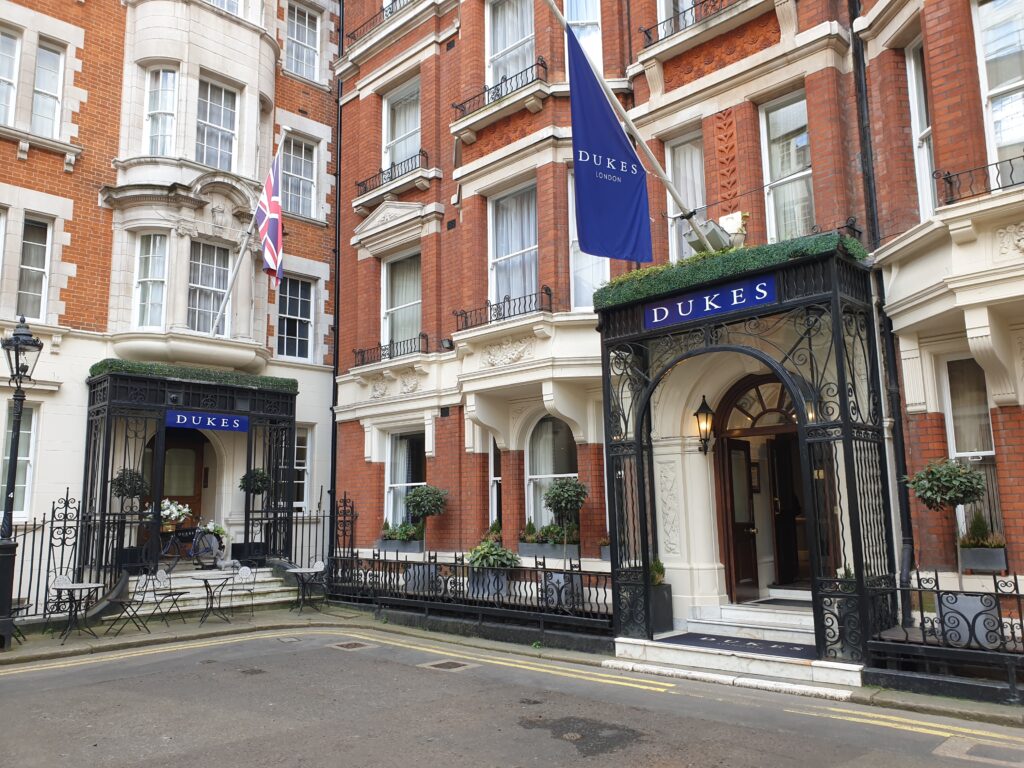 A brick building with iron cast entrance. A blue sign over the door saying "Dukes", and a flag hanging over it on a pole saying "Dukes London".