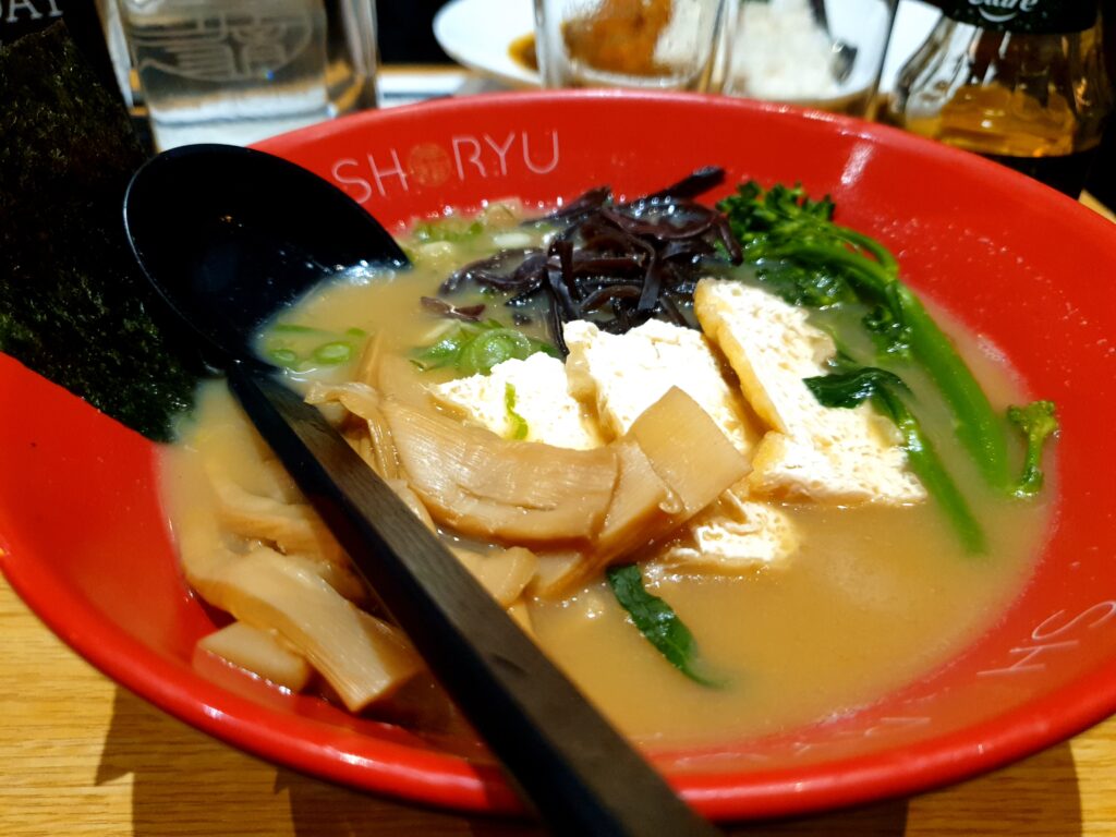 A red bowl filled wit ramen: mushrooms, spring onions tofu, broccolini, ans sweet potato.