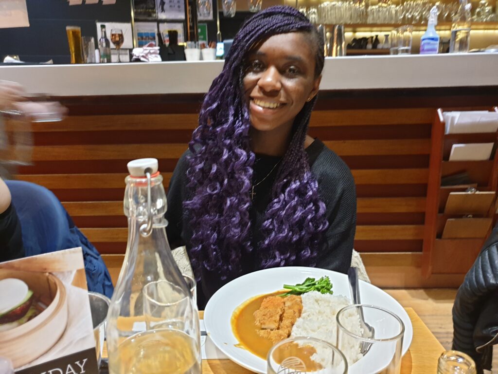 A Black woman with braided, purple hair is sitting behind a table, smiling. On the table in front of her is a plate with chicken katsu curry and rice.