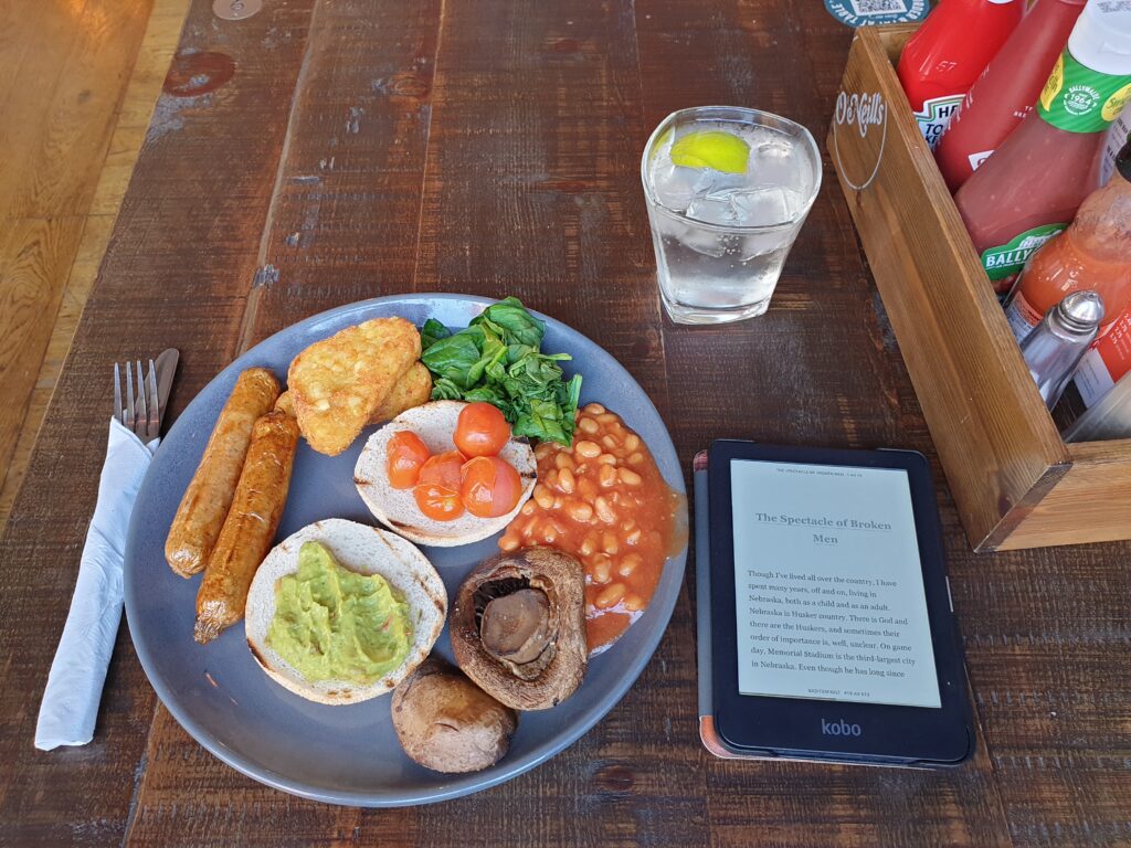 A plate with vegan sausages, half of a bap with mushed avocado, half a bap with cherry tomatoes, hash browns, spinach, baked beans, and mushrooms. A glass with fizzy water behind it, and an e-reader with an open page to the right.