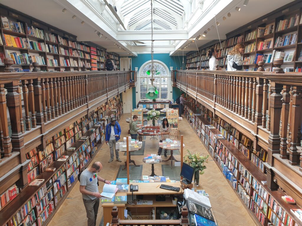 A room with walls covered with books. At the end of the room is a window with painted glass art. Flowerlike green lamps are hanging from the ceiling. There are also tables filled with books in the room.