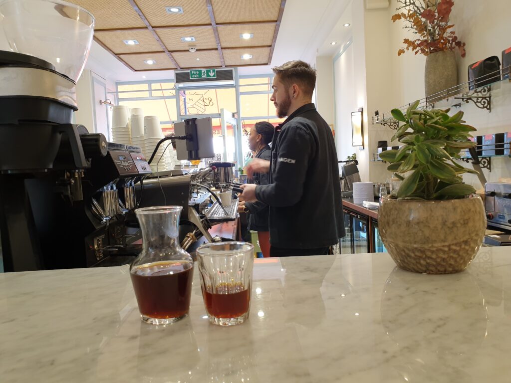 A marble counter with a carafe and a glass of coffee. To the left behind the counter you can see coffee grinders, espresso machines, and other coffee making equipment. You can also see two baristas working.