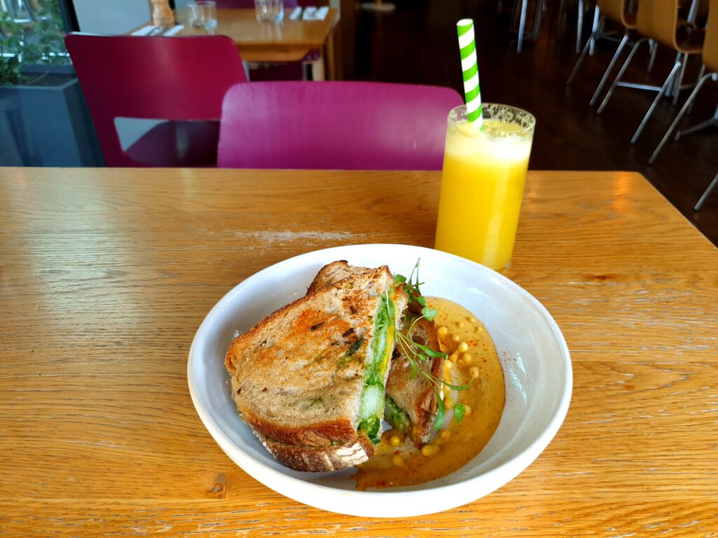 A white plate with a toasted sandwich with some green spread, and a mango pickle yoghurt. Behind it a glass of yellow pinapple, apple, tumeric, and ginger juice with a green and white striped straw.