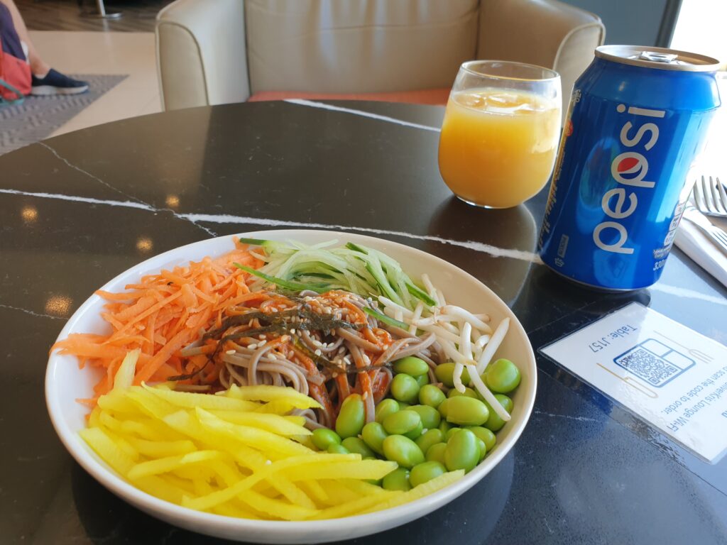 A white bowl with vegetables, noodles, and soy beans. A glass of orange juice and a can of Pepsi behind it.
