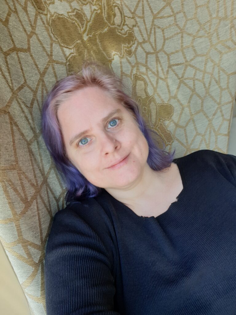 A woman with purple hair and blue eyes, and a faint smile. She's wearing a black sweater. She seems to be relaxing in a chair.