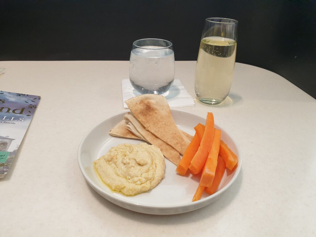 A bowl with hummus,flatbread, and carrot sticks on a table. Behind it a glass of water and a glass of bubbly wine.