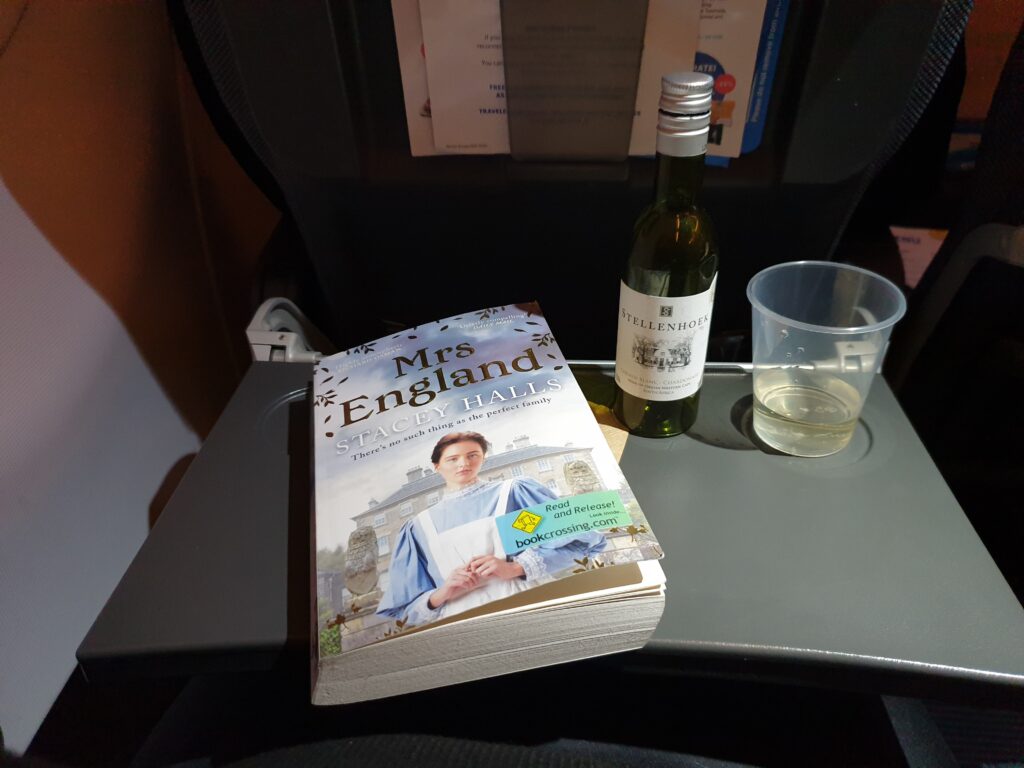 A flight seat table flipped open, on it a book to the left, and a plastic cup with white wine to the right. Between them a small, plastic white wine bootle.