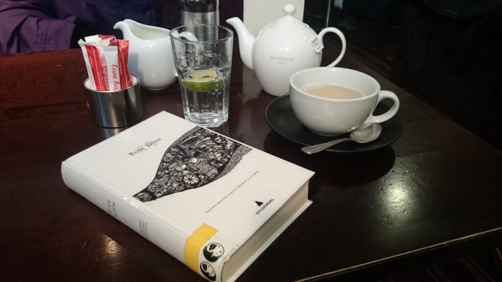 A book is on a table, behind it is a tea cup, a tea pot, and a glass of water.