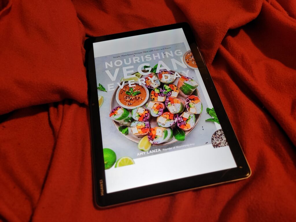 A tablet laying on a red blanket. On the screen you can see the cover of the book "Nourishing Vegan Every Day" by Amy Lanza.