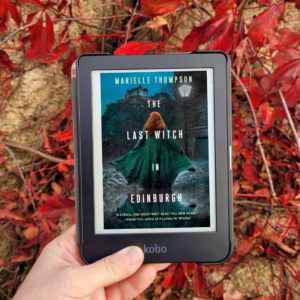 A hand holding an e-reader showing the book The Last Witch in Edinburgh. In the background is a rock covered in red leaves.